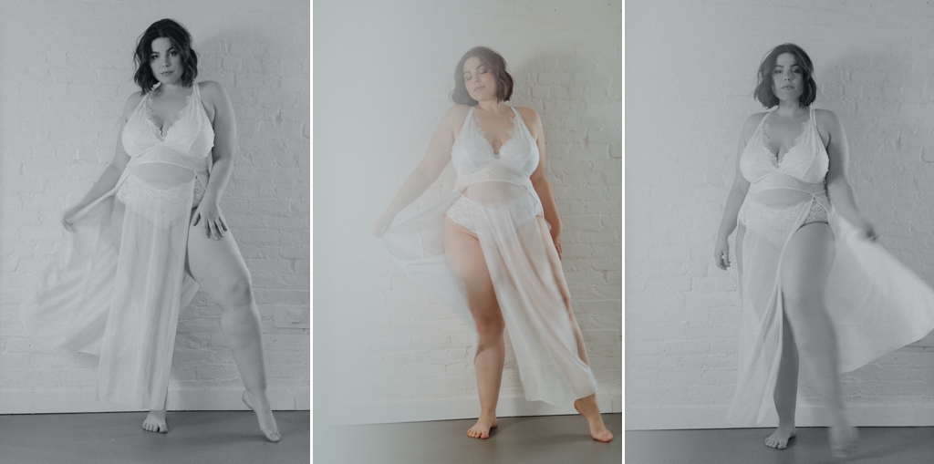 plus sized woman stands in front of a white brick wall, poses for boudoir photos in lacy two piece set and sheet dress