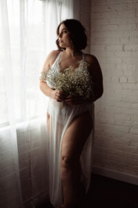 plus sized brunette stands in front of a glowing window with gauzy curtain, wears a lacy two piece set with a sheer dress
