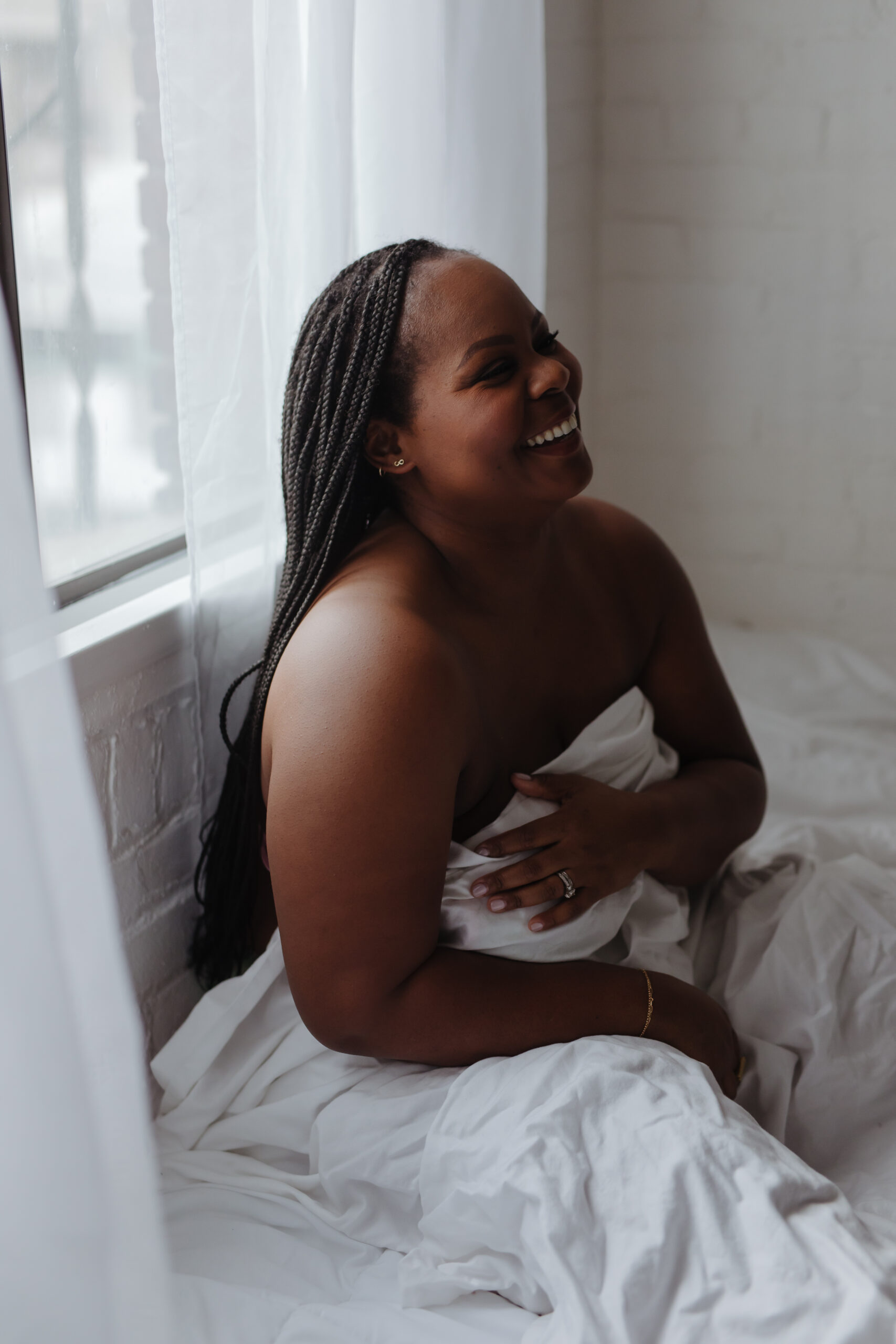 a black woman with braids laughs while wrapped in a white sheet for a boudoir photography session